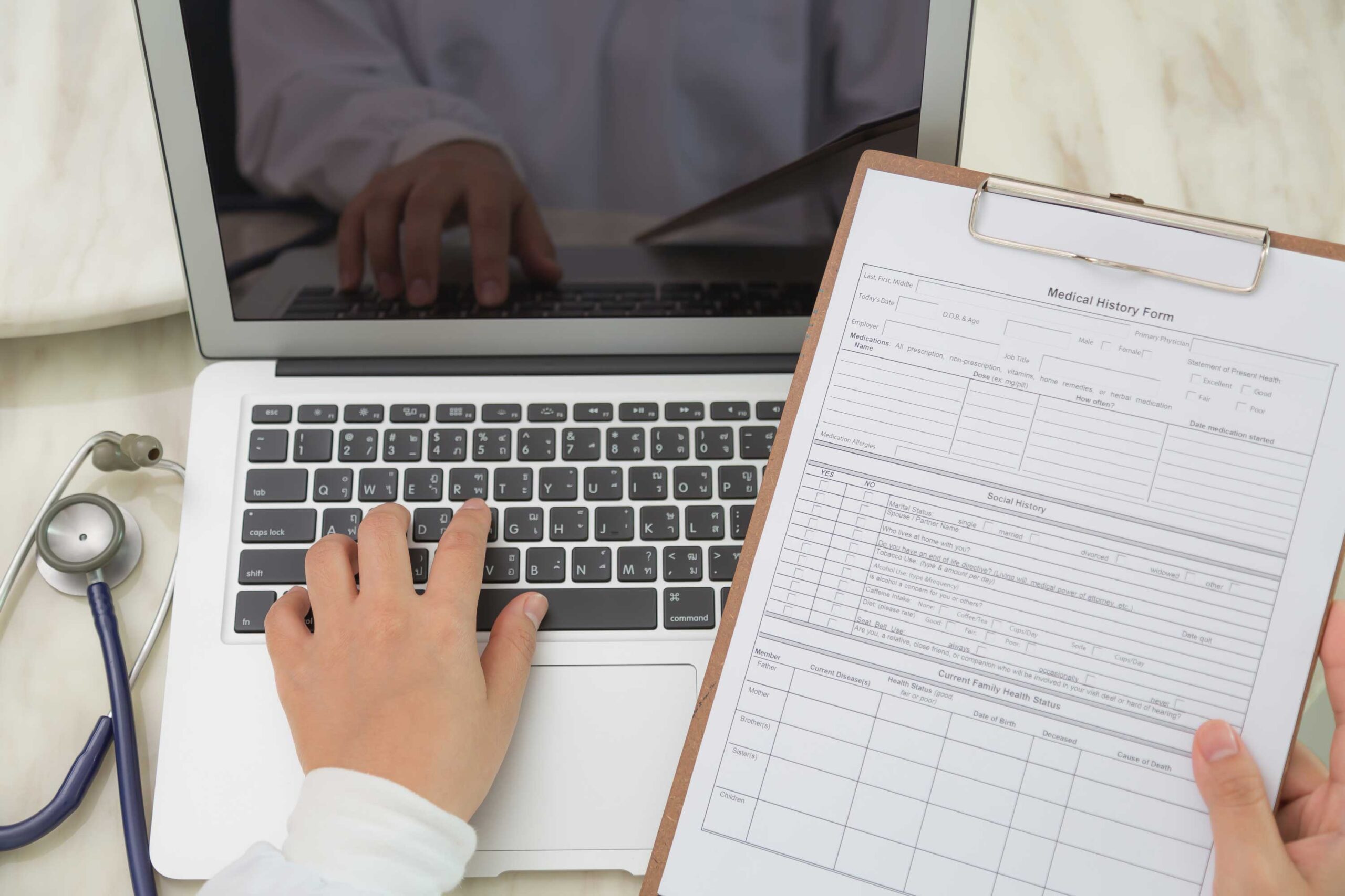 View of doctor using a laptop with a clipboard in hand